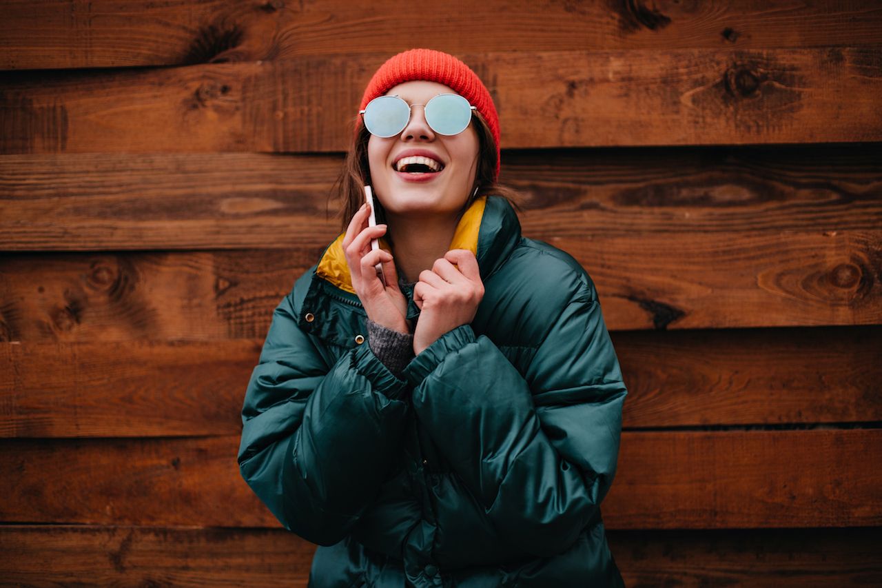 young-woman-great-mood-genuinely-laughs-while-talking-phone-girl-red-hat-emerald-color-jacket-poses-wooden-background.jpg